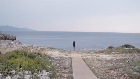 Girl-walking-on-beach-trail-and-looking-towards-sea-cliff,-wide-shot