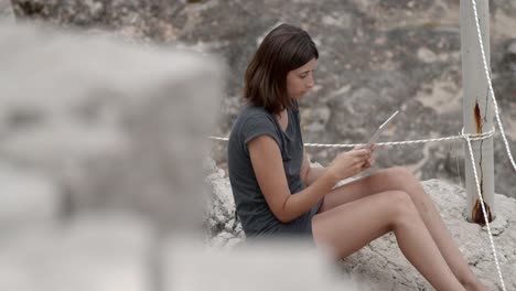 Girl-sitting-on-a-beautiful-rocky-beach-with-tablet-computer-in-her-hand