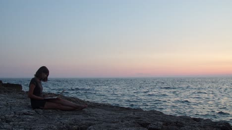 Mädchen-Sitzt-Auf-Einem-Felsen-Am-Meer-Mit-Laptop-Und-Sonnenuntergang-Im-Hintergrund,-Zeitlupe