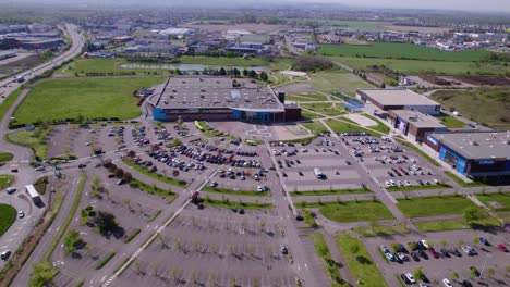 aerial-view-by-drone-of-a-shopping-center-with-a-huge-open-air-parking-lot,-parked-cars-and-cars-in-traffic,-big-road-on-the-left