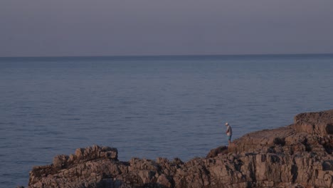 Man-with-a-hat-fishing-from-a-rock-by-beautiful-sea,-golden-hour,-copyspace