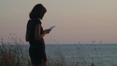 Girl-standing-on-a-cliff-by-the-sea-on-sunset,-with-tablet-computer-in-hands-wider-shot