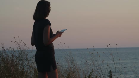 Girl-with-glasses-standing-on-a-cliff-by-the-sea-on-sunset--golden-hour,-taking-a-photo-with-tablet-computer