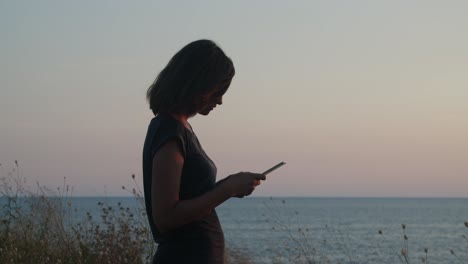 Girl-standing-on-a-cliff-by-the-sea-on-sunset--golden-hour,-with-tablet-computer-in-hands-close
