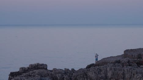 Man-fishing-from-a-rock-by-beautiful-sea,-blue-hour,-wide-shot
