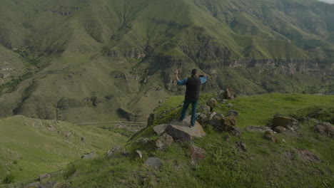 Man-Standing-On-Rocky-Mountain-Holding-Smartphone-With-Overview-Of-Mountainous-Landscape-In-Rural-Georgia