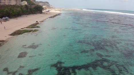 Drone-Flyover-Tropical-Melasti-Beach-Bali-Indonesia-Sobre-El-Océano-Y-La-Gente-Nadando-Y-Surfeando