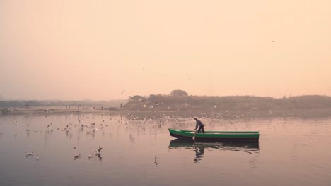 Ein-Bootsmann-Bei-Sonnenaufgang-Am-Ghat-Des-Yamuna-Flusses-Mit-Booten,-Zugvögeln,-Delhi,-Indien