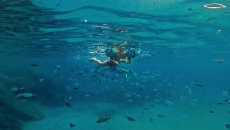 Underwater-view-of-child-swimming-with-inflatable-armbands-in-middle-of-big-school-of-fish-in-deep-blue-water