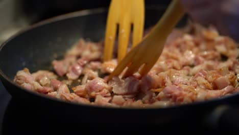 Mixing-diced-bacon-slices-in-frying-pan-as-it-sizzles,-handheld-close-up