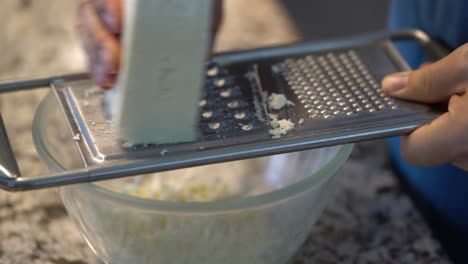 Close-up-of-hand-grating-block-of-feta-on-cheese-shredder-in-home-kitchen