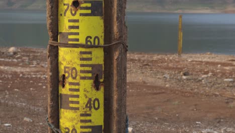 Tilting-up-shot-of-an-old-rusty-water-level-gauge-post-on-the-water-edge-with-view-of-lake,-mountain-range,-and-blue-sky-at-a-national-park
