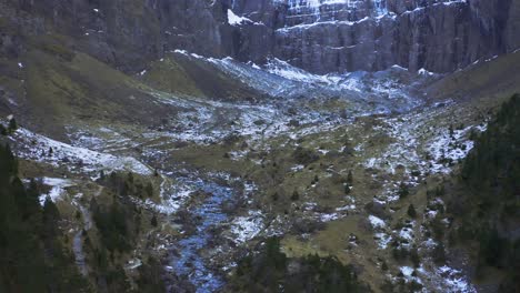 Shot-from-a-drone-of-a-mountain-cliff-with-snow-on-the-top-panning-downward-revealing-a-little-water-river-source