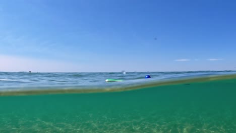 Unusual-split-underwater-view-of-two-lost-beach-tennis-paddles-and-blue-ball-floating-on-sea-water-surface