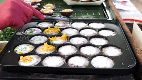 Street-food-vendor-is-preparing-Kanom-Krok-or-Thai-coconut-pudding,-a-kind-of-ancient-Thai-dessert-snack-with-choices-of-toppings-such-as-spring-onion,-corn-kernels,-taro,-etc