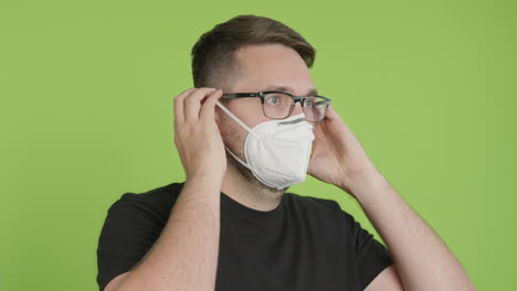 Man-in-Glasses-Puts-on-Protective-KN95-Face-Mask-While-Looking-Right-of-Camera-on-Green-Screen-Chroma-Key