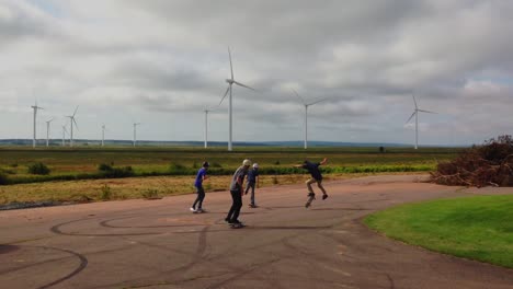 Four-kids-skate-boarding-with-backdrop-of-wind-turbines,-with-clouds