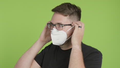 Man-in-Glasses-Puts-on-Protective-KN95-Face-Mask-While-Looking-Left-of-Camera-on-Green-Screen-Chroma-Key