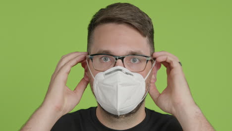Man-in-Glasses-Puts-on-Protective-KN95-Face-Mask-While-Looking-Into-Camera-on-Green-Screen-Chroma-Key-Close-Up