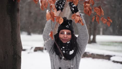 woman-in-the-winter-in-a-park-holding-hands-up-and-dropping-snow-on-her-while-smiling-with-leaves-in-front-mid-shot