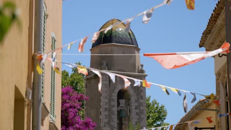 full-of-little-colored-flags-flying-in-the-wind,-tied-on-a-rope-hanging-between-two-walls-of-an-alley-with-a-church-and-its-bell-in-the-background,-sunny-weather,-blue-sky