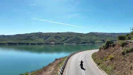 Motociclista-Montando-Solo-En-Una-Carretera-Vacía-Con-Vistas-A-Un-Hermoso-Paisaje-Con-Un-Lago-Azul