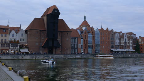 riverside-view-of-crane-Zuraw-at-Gdansk-Poland-historical-building-,-boat-passes-by-and-people-walking-by-the-shore-,-day-shot