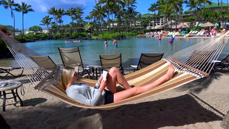 Young-blonde-woman-relaxing-on-hammock-at-resort-hotel-pool-and-swimming-lagoon-in-Poipu-Beach,-Kauai,-Hawaii,-4k,-static,-reading-a-book