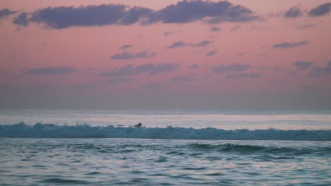 Ein-Einsamer-Surfer,-Der-Auf-Seinem-Brett-Liegt-Und-Einen-Frostigen,-Wolkigen-Rosa-Sonnenuntergang-Beobachtet