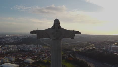 Drone-Hace-Un-Círculo-Completo-Alrededor-De-La-Poderosa-Estatua-De-Cristo-Rei-Jesucristo-Con-Una-Vista-Espectacular-En-El-Horizonte-De-Lisboa-Puente-Tejo-En-Un-Día-De-Invierno-Con-Sol-Desde-Atrás