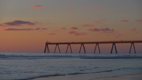 Ein-Verbrannter-Orangefarbener-Sonnenuntergang-Am-Strand-Mit-Hereinrollenden-Wellen-Und-Einem-Langen-Pier