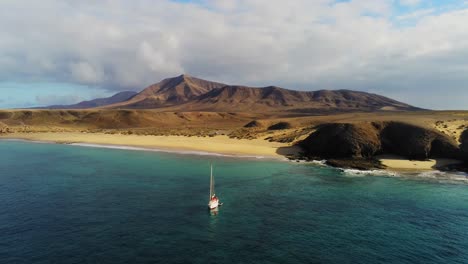 Lonely-sailboat-near-majestic-coastline-of-Lanzarote-with-mountain-in-background,-aerial-view