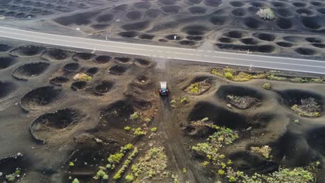 Vehicle-leaving-dirt-road-to-paved-highway-in-crater-farm-with-grapes-in-Lanzarote,-aerial-view