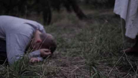 Man-in-robe-walks-up-to-teen-boy-crying-on-ground-in-forest