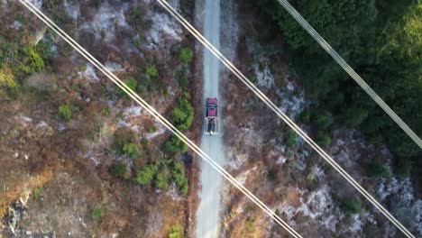 Top-view-drone-shot-following-a-pickup-truck-on-a-gravel-road-driving-into-the-forest-below-powerlines