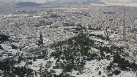 Aerial-drone-view-over-a-park-towards-snowy-cityscape-of-Athens,-winter-in-Greece