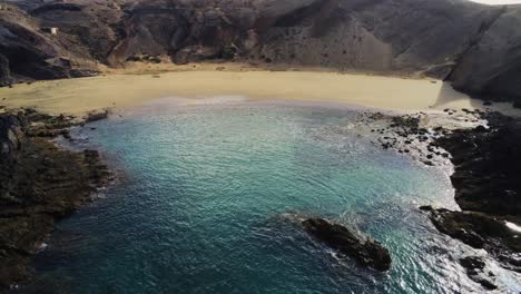 Sandy-beach-near-rocky-terrain-and-blue-ocean-water-in-Lanzarote-island,-aerial-view