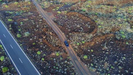 Dark-car-driving-on-gravel-rural-road-towards-highway-in-Lanzarote,-aerial-view