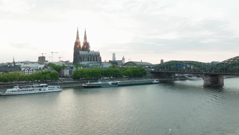 Drone-films-a-ship-on-the-Rhine-in,-the-drone-moves-up-and-you-can-see-the-Cologne-Cathedral-in-the-sunset,-a-train-drives-on-the-Hohenzoller-Bridge-over-the-Rhine