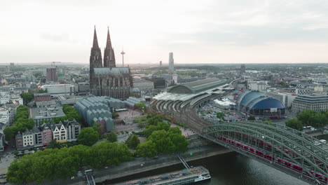 Drohne-Filmt-Den-Kölner-Dom,-Den-Kölner-Hauptbahnhof,-Das-Kölner-Theater-Im-Sonnenuntergang,-Die-Drohne-Fährt-Herunter-Und-Der-Rhein-Mit-Der-Hohenzollerbrücke-Kommt-Ins-Bild