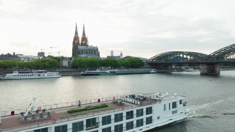 Drone-films-a-ship-on-the-Rhine-in-slow-motion,-the-drone-moves-up-and-you-can-see-the-Cologne-Cathedral-in-the-sunset,-a-train-drives-on-the-Hohenzoller-Bridge-over-the-Rhine