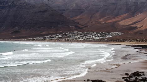 Ocean-waves-rolling-over-sandy-beach-with-small-town-in-horizon-near-massive-mountain,-static-slow-motion-view