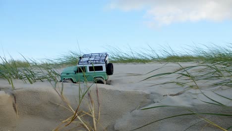 Small-remote-control-jeep-car-toy-drive-on-sandy-dunes-with-coastal-plants,-static-view