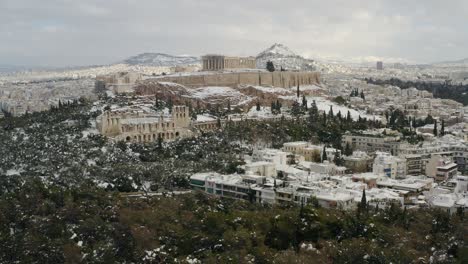 Aerial-view-of-the-snow-covered-Acropolis-hill,-blizzard-aftermath,-in-cloudy-Athens---tracking,-drone-shot