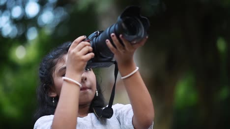 A-cute-little-Asian-girl-is-taking-photograph-of-nature-and-trees-with-a-Digital-SLR-camera-like-a-professional-photographer