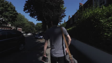 Moving-shot-following-caucasian-man,-wearing-grey-t-shirt-with-camera-bag-slung-over-one-shoulder-carrying-camera,-walking-down-street-in-North-London-Highgate-UK-day-time-sunny