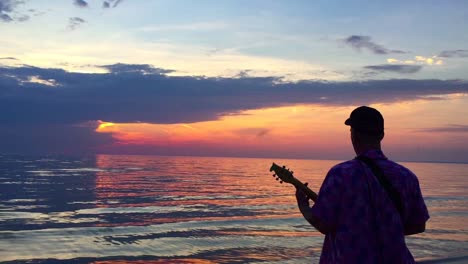 Handheld-on-man-with-back-to-camera-playing-a-busker-toy-guitar-to-the-setting-sun-on-a-beautiful-evening-at-Georgian-Bay