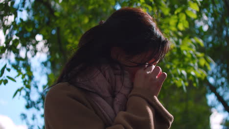 Shot-of-woman-crying,-hiding-face-in-hands-while-visiting-a-park-surrounded-by-green-trees-during-morning-time