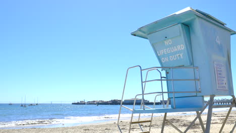 Sitting-on-the-sandy-beach-watching-the-blue-Pacific-Ocean-waves-against-the-shore-with-an-empty-lifeguard-tower-on-a-sunny-clear-day-in-Santa-Barbara,-California