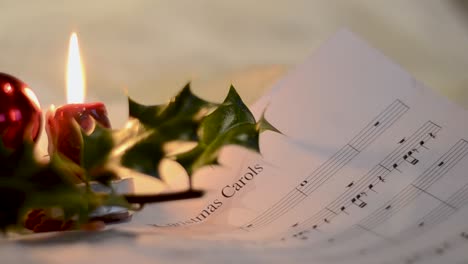 A-close-well-focused-shot-of-a-white-sheet-contaning-musical-notations-for-christmas-carol-kept-beside-a-chistmas-holly-plant-and-a-burning-small-candle